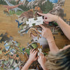 Two people working on a dinosaur-themed puzzle on a wooden table.